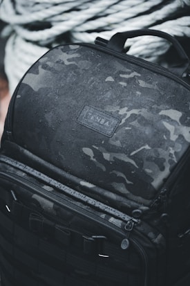 A rugged black backpack with a camouflage pattern is covered in water droplets, indicating it is water-resistant. The backpack features multiple zippers and handles, and it is positioned against a backdrop of thick, coiled ropes.