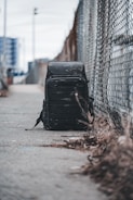 A black backpack rests on a concrete sidewalk beside a chain-link fence. The background features an urban setting with blurred skyscrapers, creating a sense of isolation in a city environment. The ground is scattered with dried grass, and the sky appears overcast.