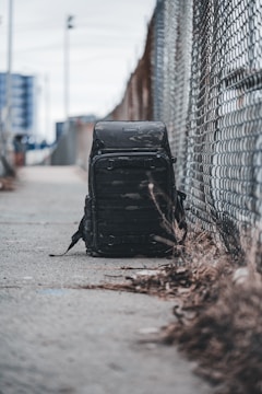 A black backpack rests on a concrete sidewalk beside a chain-link fence. The background features an urban setting with blurred skyscrapers, creating a sense of isolation in a city environment. The ground is scattered with dried grass, and the sky appears overcast.