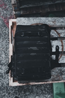 A black tactical backpack with multiple compartments is placed on a wooden pallet. The background consists of a concrete surface and other wooden pallets stacked nearby.