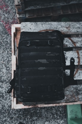 A black tactical backpack with multiple compartments is placed on a wooden pallet. The background consists of a concrete surface and other wooden pallets stacked nearby.