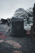 A rugged backpack sits on a rough, gravelly surface in an outdoor setting. Large coiled ropes are piled in the background, suggesting a maritime or industrial environment.