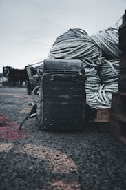 A rugged backpack sits on a rough, gravelly surface in an outdoor setting. Large coiled ropes are piled in the background, suggesting a maritime or industrial environment.