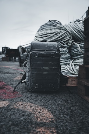 A rugged backpack sits on a rough, gravelly surface in an outdoor setting. Large coiled ropes are piled in the background, suggesting a maritime or industrial environment.