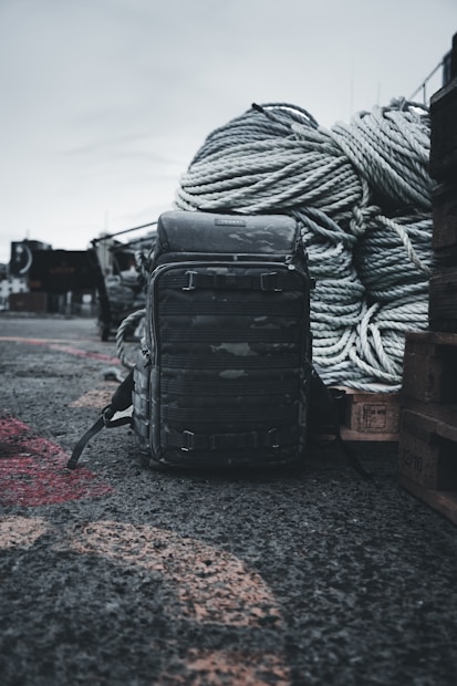 A rugged backpack sits on a rough, gravelly surface in an outdoor setting. Large coiled ropes are piled in the background, suggesting a maritime or industrial environment.