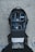 A black camera backpack on a concrete surface, photographed from above. The backpack is open, revealing various camera equipment inside, including several lenses, a camera body, a flash, and other accessories neatly organized in padded compartments. A pair of brown shoes and part of a person’s legs are visible at the bottom edge of the image.