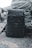 A dark camouflage-patterned backpack is placed on a rough concrete surface, with large coiled ropes stacked in the background. The setting appears industrial and utilitarian, with muted colors and textures that convey a sense of ruggedness.