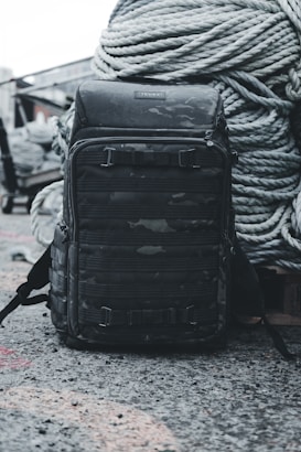 A dark camouflage-patterned backpack is placed on a rough concrete surface, with large coiled ropes stacked in the background. The setting appears industrial and utilitarian, with muted colors and textures that convey a sense of ruggedness.