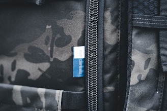 A close-up view of a backpack featuring a camouflage pattern with a visible zipper. Attached to the zipper area is a small blue tag with the words 'NEVER COMPROMISE' written on it.