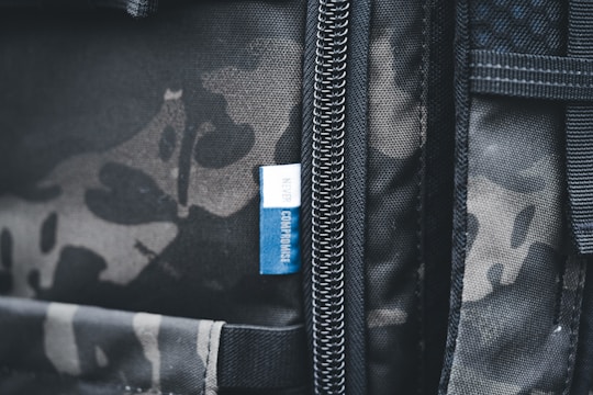 A close-up view of a backpack featuring a camouflage pattern with a visible zipper. Attached to the zipper area is a small blue tag with the words 'NEVER COMPROMISE' written on it.