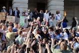 A large group of people are gathered on the steps of a building, seemingly participating in a protest or demonstration. Many individuals hold up signs with messages such as 'Bill Lee the shame of Tennessee,' 'GOP guns people shame,' and 'Gays against guns.' Several participants are taking photos or videos with their phones, capturing the event. A person is seen speaking through a megaphone, while others around them appear engaged and attentive. The gathering includes a diverse mix of people, including men, women, and elderly individuals.