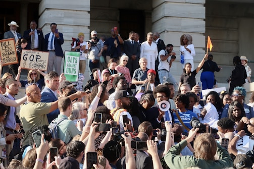 A large group of people are gathered on the steps of a building, seemingly participating in a protest or demonstration. Many individuals hold up signs with messages such as 'Bill Lee the shame of Tennessee,' 'GOP guns people shame,' and 'Gays against guns.' Several participants are taking photos or videos with their phones, capturing the event. A person is seen speaking through a megaphone, while others around them appear engaged and attentive. The gathering includes a diverse mix of people, including men, women, and elderly individuals.
