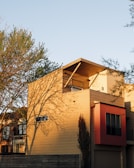 A modern house with a flat roof and a rectangular structure, featuring a combination of beige and red exterior panels. The building is surrounded by trees casting distinct shadows on the facade, with sunlight creating a warm glow.