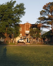A brick house with tall trees on either side, a white SUV parked in the driveway, and a well-manicured lawn in the foreground. The sunlight casts long shadows, creating a warm and serene atmosphere. A decorative metal fence and gate enclose the front yard.