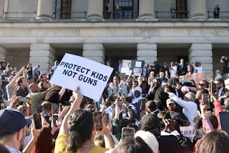 A large group of people gathered in front of a building with columns, holding signs related to gun control advocacy. The central sign has the message 'PROTECT KIDS NOT GUNS.' Many participants are taking photos or filming the event with their phones. The atmosphere is energetic and crowded, with people of various backgrounds.