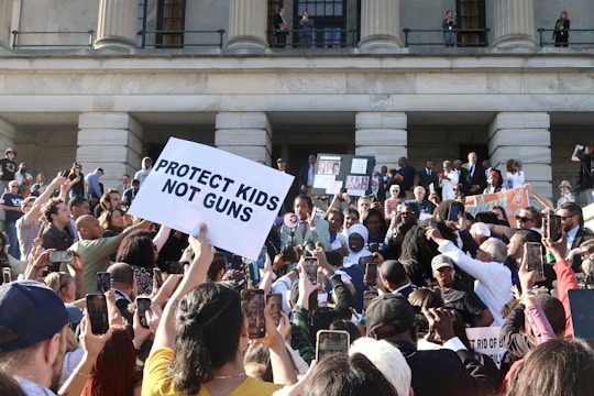 A large group of people gathered in front of a building with columns, holding signs related to gun control advocacy. The central sign has the message 'PROTECT KIDS NOT GUNS.' Many participants are taking photos or filming the event with their phones. The atmosphere is energetic and crowded, with people of various backgrounds.