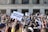 A large group of people gathered in front of a building with columns, holding signs related to gun control advocacy. The central sign has the message 'PROTECT KIDS NOT GUNS.' Many participants are taking photos or filming the event with their phones. The atmosphere is energetic and crowded, with people of various backgrounds.