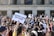 A large group of people gathered in front of a building with columns, holding signs related to gun control advocacy. The central sign has the message 'PROTECT KIDS NOT GUNS.' Many participants are taking photos or filming the event with their phones. The atmosphere is energetic and crowded, with people of various backgrounds.