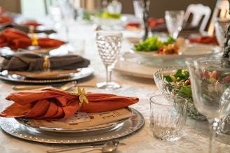 A dining table is elegantly set with glassware, plates, and intricately folded napkins with decorative rings. Plates are arranged with menus or placeholders, and bowls of fresh salad with greens, tomatoes, and other vegetables are positioned around the table. Crystal glasses and silverware add a touch of sophistication.