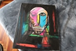 An abstract painting featuring a stylized human face composed of geometric shapes and bold lines. The face includes one prominently defined eye, an angular nose, and a mouth with simplified features. The background is a mix of vibrant colors creating a dynamic contrast.