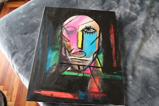An abstract painting featuring a stylized human face composed of geometric shapes and bold lines. The face includes one prominently defined eye, an angular nose, and a mouth with simplified features. The background is a mix of vibrant colors creating a dynamic contrast.