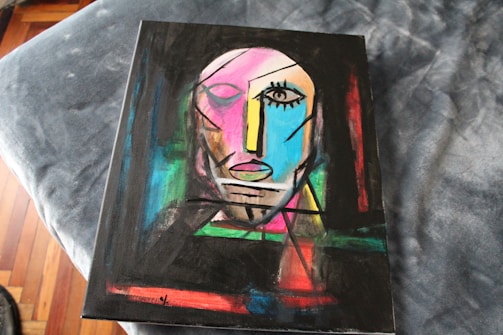 An abstract painting featuring a stylized human face composed of geometric shapes and bold lines. The face includes one prominently defined eye, an angular nose, and a mouth with simplified features. The background is a mix of vibrant colors creating a dynamic contrast.