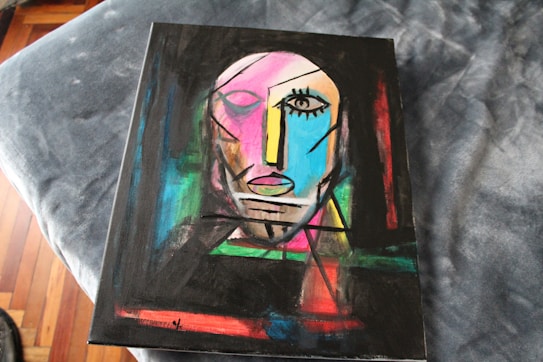 An abstract painting featuring a stylized human face composed of geometric shapes and bold lines. The face includes one prominently defined eye, an angular nose, and a mouth with simplified features. The background is a mix of vibrant colors creating a dynamic contrast.