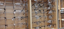 A display of multiple pairs of eyeglasses arranged neatly on metal racks against a wooden background. The eyeglass frames vary in style and color, including round, rectangular, and square shapes. Price tags are attached to each pair, indicating they're for sale.