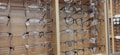A display of multiple pairs of eyeglasses arranged neatly on metal racks against a wooden background. The eyeglass frames vary in style and color, including round, rectangular, and square shapes. Price tags are attached to each pair, indicating they're for sale.