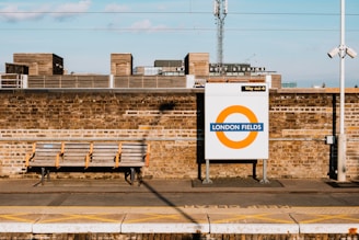 A train station platform features a brick wall with a signage for London Fields with a circular orange and blue design. Above, there are industrial-style buildings with a visible sign for Netil Studios. A bench is placed on the platform, providing seating.