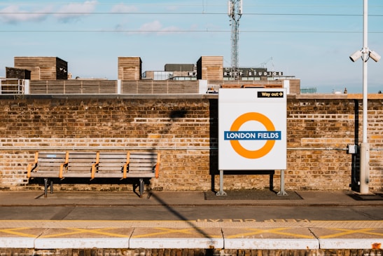 A train station platform features a brick wall with a signage for London Fields with a circular orange and blue design. Above, there are industrial-style buildings with a visible sign for Netil Studios. A bench is placed on the platform, providing seating.