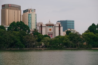 A cityscape featuring several tall buildings including a prominent one with the name 'Seabank' on top. The foreground shows a body of water bordered by lush green trees. The overall scene conveys a blend of urban architecture and natural scenery.