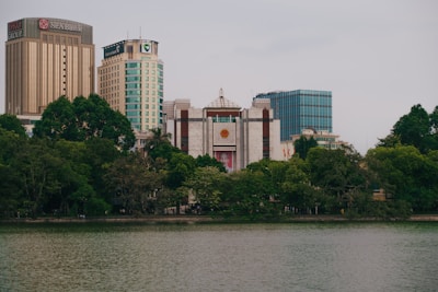 A cityscape featuring several tall buildings including a prominent one with the name 'Seabank' on top. The foreground shows a body of water bordered by lush green trees. The overall scene conveys a blend of urban architecture and natural scenery.