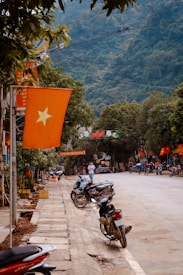 A street view featuring multiple parked motorcycles along a sidewalk, with a row of orange flags displaying a yellow star on the left side. The background reveals lush green hills, and people can be seen walking and bicycling on the street. The scene is lined with trees, adding to the greenery.
