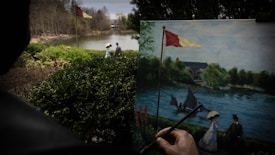 A painting in progress shows a lakeside scene with trees and a small sailboat. The painting depicts a couple dressed in vintage clothing, with the woman holding an umbrella, walking by the lake. In the foreground, bushes and trees frame the view of the river setting.