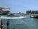 A bustling harbor scene showcases a large cruise ship docked alongside smaller boats navigating the water. The iconic Sydney Opera House stands prominently in the background against a backdrop of clear blue sky. High-rise buildings are visible adjacent to the water, while the view captures a lively atmosphere with movement on the water.