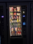 A vending machine filled with various snacks and beverages. The upper rows contain packages of chips, cookies, and snacks, while the lower sections are stocked with bottles and cans of drinks. A keypad and payment interface are visible on the right side.
