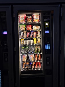 A vending machine filled with various snacks and beverages. The upper rows contain packages of chips, cookies, and snacks, while the lower sections are stocked with bottles and cans of drinks. A keypad and payment interface are visible on the right side.