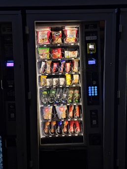 A vending machine filled with various snacks and beverages. The upper rows contain packages of chips, cookies, and snacks, while the lower sections are stocked with bottles and cans of drinks. A keypad and payment interface are visible on the right side.