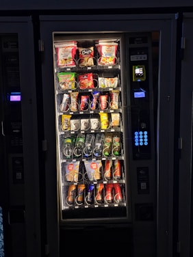A vending machine filled with various snacks and beverages. The upper rows contain packages of chips, cookies, and snacks, while the lower sections are stocked with bottles and cans of drinks. A keypad and payment interface are visible on the right side.