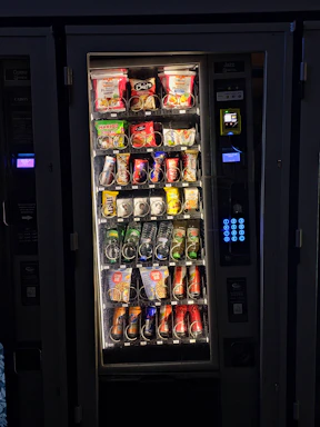 A vending machine filled with various snacks and beverages. The upper rows contain packages of chips, cookies, and snacks, while the lower sections are stocked with bottles and cans of drinks. A keypad and payment interface are visible on the right side.