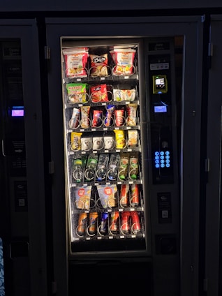 A vending machine filled with various snacks and beverages. The upper rows contain packages of chips, cookies, and snacks, while the lower sections are stocked with bottles and cans of drinks. A keypad and payment interface are visible on the right side.