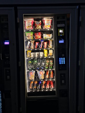 A vending machine filled with various snacks and beverages. The upper rows contain packages of chips, cookies, and snacks, while the lower sections are stocked with bottles and cans of drinks. A keypad and payment interface are visible on the right side.
