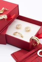 A red jewelry box contains ornate golden earrings and a ring. The box is open, revealing a white interior that highlights the jewelry. There are additional golden rings placed nearby, adding to the luxurious display.
