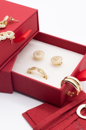 A red jewelry box contains ornate golden earrings and a ring. The box is open, revealing a white interior that highlights the jewelry. There are additional golden rings placed nearby, adding to the luxurious display.