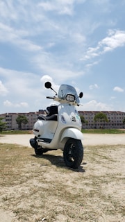 A white scooter is parked on an open sandy area with a background of apartment buildings under a partly cloudy blue sky.