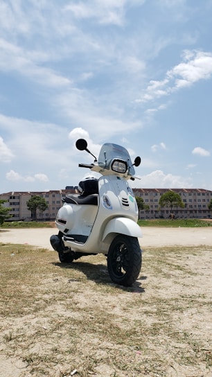 A white scooter is parked on an open sandy area with a background of apartment buildings under a partly cloudy blue sky.