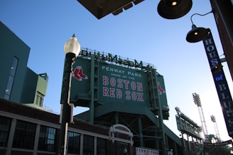 A view of Fenway Park with a large green scoreboard displaying the words 'Home of the Boston Red Sox' along with two red socks symbols. Surrounding buildings and a streetlamp are visible, with a clear blue sky in the background.