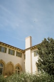 A Mediterranean-style building is depicted with light stucco walls and green shutters. The house features arched windows and a prominent chimney. In the foreground, lush greenery and flowering shrubs are visible. The backdrop is a clear blue sky with a small bird in flight.
