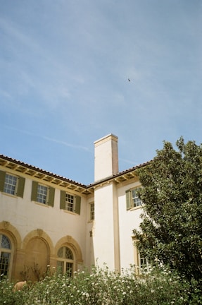 A Mediterranean-style building is depicted with light stucco walls and green shutters. The house features arched windows and a prominent chimney. In the foreground, lush greenery and flowering shrubs are visible. The backdrop is a clear blue sky with a small bird in flight.